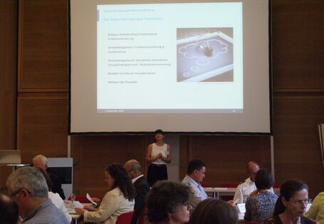 Vortragende im weißen sommerlichen Shirt steht vor der Präsentationsleinwand und spricht zu den Teilnehmenden. Der Raum ist verdunkelt. Die Teilnehmenden schauen nachdenklich auf den Papieren in ihren Händen. Sie sitzen an Gruppenarbeitstischen.