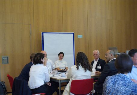 Teilnehmende sitzen an einem Gruppenarbeitstisch und hören einer Teilnehmerin zu. Im Hintergrund steht eine Pinwand mit einem großen leeren weißen Blatt bespannt. 