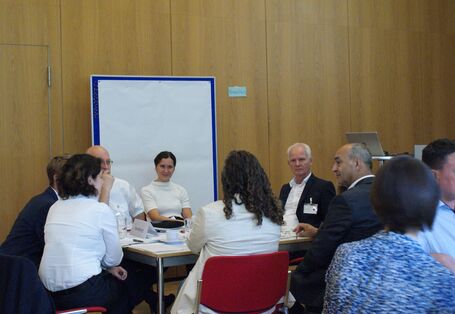 Teilnehmende sitzen an einem Gruppenarbeitstisch und hören einer Teilnehmerin zu. Im Hintergrund steht eine Pinwand mit einem großen leeren weißen Blatt bespannt.