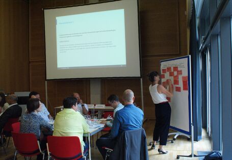 Teilnehmende präsentiert an Moderationswand mit roten Moderationskarten. Die anderen Teilnehmenden sitzen nachdenklich am Gruppenarbeitstisch. Im Hintergrund ist eine Präsentation eingblendet.