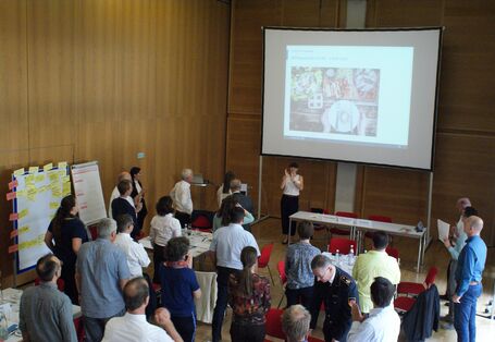 Alle Teilnehmenden stehen im Raum und folgen der Präsentation einer Teilnehmerin, die vor der Leinwand steht. Manche Teilnehmende applaudieren. Teile der Präsentationswände mit den farbigen Moderationskarten stehen am Rand verteilt