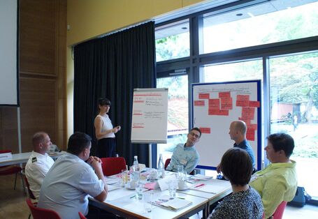 Teilnehmende sitzen an ihrem Gruppenarbeitstisch in aufgelockerter Atmosphäre vor einer großen Fensterfront, die den Blick ins Grüne freigibt. Zwischen Fensterfront und Gruppenarbeitstisch stehen zwei Präsentationswände mit roten Moderationskarten bestückt und beschriftet.