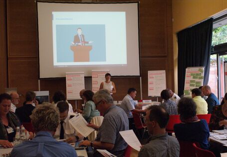 Teilnehmende sitzen in lockerer Atmosphäre an ihren Tischen und halten Papiere in der Hand, die sie sichten. Im Hintergrund stehen die Präsentationswände an den Rändern verteilt. Eine Teilnehmerin spricht vor der Leinwand zu allen anderen Teilnehmenden.