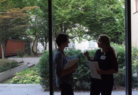Zwei Teilnehmende stehen vor der Fensterfront und tauschen sich miteinander aus. Sie halten Unterlagen in den Händen sowie Stifte. Die Fensterfront gibt einen Blick frei ins Grüne.