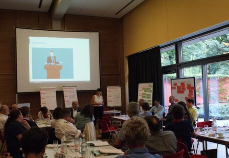 Die Teilnehmenden sitzen in lockerer Atmosphäre an ihren Arbeitstischen und hören einer Teilnehmerin zu, die am Kopfende des Raumes steht vor den aufgereihten Moderationswänden, die mit den farbigen Moderationskarten bestückt und beschriftet sind. Die große Fensterfront rechts davon gibt den Blick frei ins Grüne.