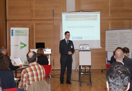 Vortragender steht und faltet die Hände. Im Hintergrund stehen Moderationswände, Veranstaltungsaufsteller und eine Leinwand. Im Vordergrund sitzen Zuhörer.