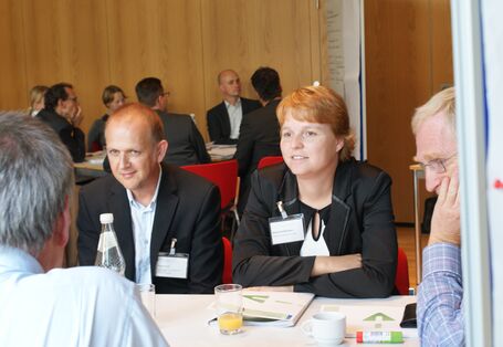 Zwei Teilnehmende sitzen nebeneinander am Gruppenarbeitstisch und hören anderen Teilnehmern zu. Im Hintergrund sitzen andere Teilnehmer am anderen Gruppenarbeitisch. Neben diesem Gruppenarbeitstisch steht eine Moderationswand.