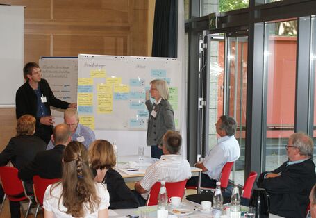 Zwei Teilnehmer stehen an Moderationswand im Dialog miteinander. An der Moderationswand hängen farbige Moderationskarten. Auf der rechten Seite steht die Tür von der Fensterfront offen. Zuhörer sitzen im Vordergrund an Gruppenarbeitstischen.