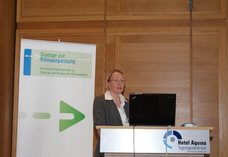 Vortragende steht am Rednerpult vor mit Holz vertäfelter Wand. Im Vordergrund steht am Rednerpult der Schriftzug des Veranstaltungsortes Tagungszentrum Hotel Aquino. Im Hintergrund steht der Veranstaltungsaufsteller Dialoge zur Klimaanpassung.