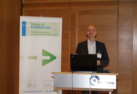 Vortragender steht am Rednerpult vor mit Holz vertäfelter Wand. Das Rednerpult trägt vorne den Schriftzug Hotel Aquino Tagungszentrum. Im Hintergrund steht der Veranstaltungsaufsteller Dialoge zur Klimaanpassung.