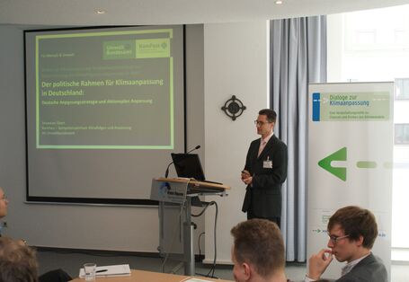 Vortragender steht am Rednerpult. Im Hintergrund sieht man die Präsentation auf Projektionswand und Veranstaltungsaufsteller.