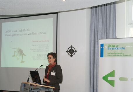 Vortragende steht am Rednerpult und trägt vor. Im Hintergrund sieht man den Vortrag auf der Projektionswand und den Veranstaltungsaufsteller.