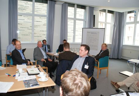 Teilnehmer sitzen an Gruppenarbeitstischen und lachen. Im Hintergrund steht eine Moderationswand vor der Fensterfront.