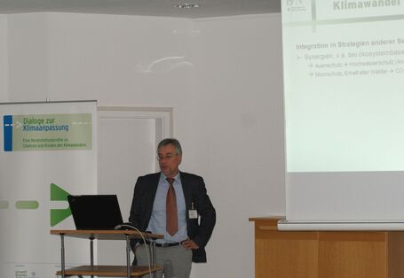 Ein Teilnehmer hält seinen Vortrag am geöffneten Laptop am Rednerpult. Präsentationsfolie wird im Hintergrund auf weiße Wand projiziert zum Thema: Integration von Strategien anderer Sektoren.