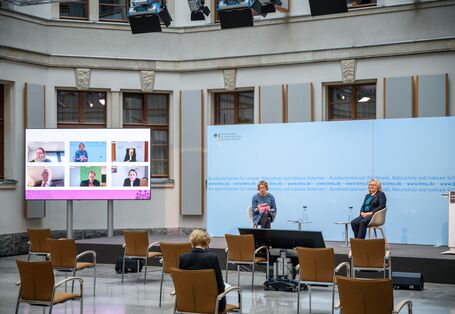 In dem hellen historischen Innenhof sitzen 2 Frauen auf Schalenstühlen auf dem Podium vor einem blauem Hintergrund. Links davon steht ein großer flacher Bildschirm, der die zugeschalteten Konferenzteilnehmenden zeigt. Vor dem Podium stehen leere Stühle. Auf einem Stuhl beugt sich eine Teilnehmerin über Ihre Unterlagen.