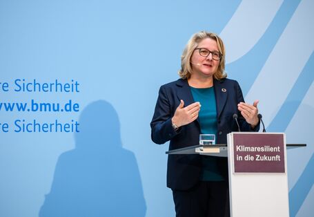 Die Umweltministerin steht am Rednerpult vor blauem Hintergrund und trägt gestikulierend vor.