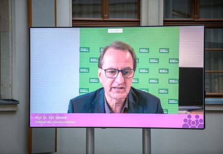 Auf einem flachen Bildschirm im hellen historischen Innenhof ist im Großformat zu sehen wie der Präsident des Umweltbundesamts einen Beitrag spricht.