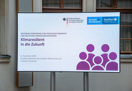 Der Blick geht auf das Konferenzlogo, welches sich im hellen historischen Innenhof auf dem großen flachen Bildschirm nahe zeigt
