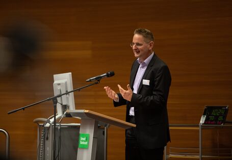 Ein Teilnehmender spricht am Stehpult in das Mikrofon. Er gestikuliert mit seinen Händen dazu und trägt einen dunklem Anzug mit hellem Hemd unter dem Jacket.