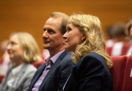 Zwei Teilnehmende sitzen im Publikum und schauen interessiert in Richtung Bühne und tauschen sich dabei aus.