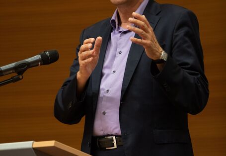 Ein Vortragender spricht gestikulierend ins Mikrofon. Er steht an einem Rednerpult und trägt einen dunklen Anzug mit einem zartlila Hemd unter dem Jacket.
