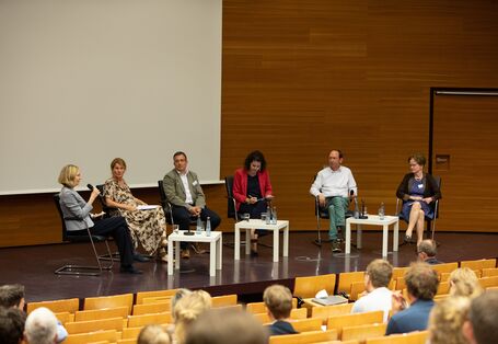 6 Podiumsteilnehmende sitzen auf der Bühne im Halbkreis vor hellen niedrigen Tischen. Auf den Tischen stehen Getränke. Eine Frau spricht in ein Mikrofon.