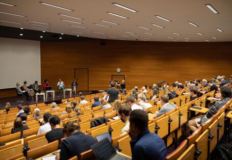 Der Blick geht über das Publikum hinweg zu der Bühne, wo die Podiumsteilnehmden vor den niedrigen hellen Tischen sitzen. Ein Teilnehmender spricht in das Handmikrofon.