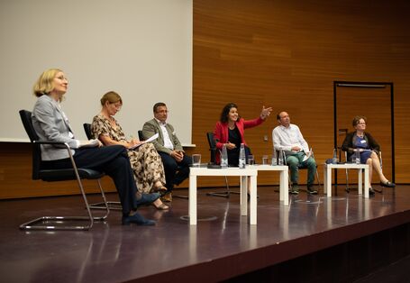 6 Podiumsteilnehmende sitzen auf der Bühne im Halbkreis vor hellen niedrigen Tischen und tauschen sich gestikulierend mit dem Publikum aus. Eine Frau im roten Sakko zeigt mit gestrecktem Arm in Richtung Publikum.
