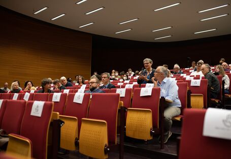 Der Blick geht von der Bühne schräg nach vorne auf das Publikum. Dort spricht eine Teilnehmende gestikulierend in ein Handmikrofon. Das Publikum hört interessiert zu.