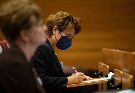 Eine Teilnehmende sitzt im Publikum und schreibt am Klapptisch in ihren Notizen.Sie trägt eine blaue FFP2 Maske.