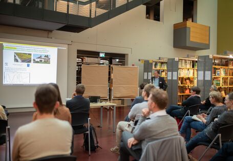 Der Blick geht über die sitzenden Workshopteilnehmenden hinüber nach Vorne auf die Leinwand. Ein Vortragender zeigt auf seine Präsentation auf der Leinwand.