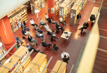 Der Blick geht von oben auf die sitzenden Worshopteilnehmenden und öffnet damit den Blick auf den Ort des Workshops: eine Bibliothek.