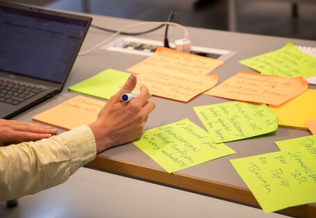 Der Blick geht auf die schreibende Hand eines Workshopteilnehmenden vor aufgeklapptem Notebook. Auf dem Tisch liegen verstreut beschriftete gelbe Moderationskarten