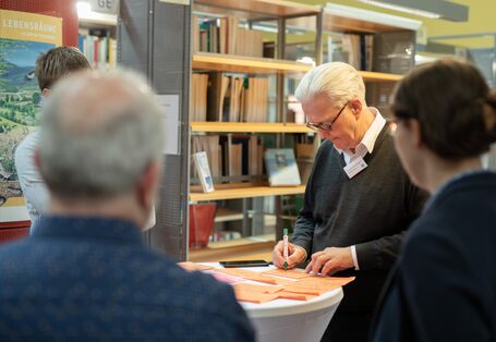 Workshopteilnehmende stehen um einen Stehtisch. Ein Teilnehmender beschriftet orangefarbene Moderationskarten.
