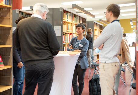 Workshopteilnehmende stehen um einen Stehtisch und tauschen sich aus. Der Blick geht auf eine Teilnehmerin mit verschränkten Armen.