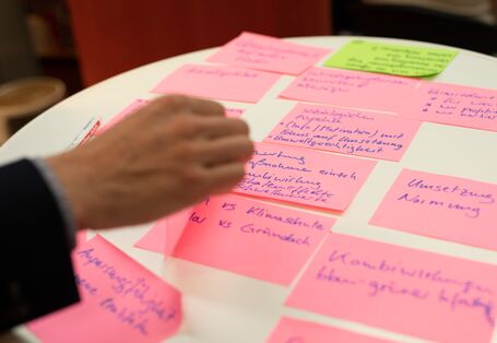 Eine Hand eines Workshopteilnehmenden sortiert rosafarbene Moderationskarten. Die Morderationskarten sind beschriftet und liegen auf einem weißen runden Tisch.