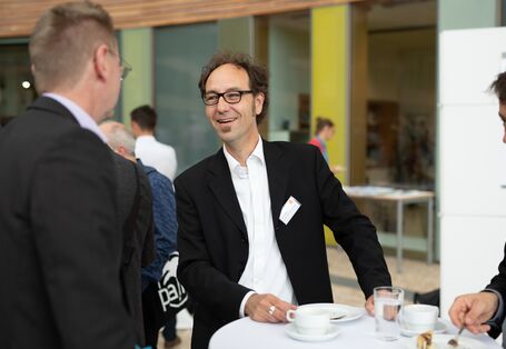 Teilnehmende in dunkler Kleidung unterhalten sich im Atrium am Veranstaltungsort vor dem Hintergrund einer mit Holz durchsetzten und verglasten Front.