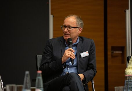 Ein Teilnehmender sitzt auf dem Podium und spricht in das Mikrofon in Richtung der anderen Podiumsteilnehmenden. Er hält das Mikrofon in der Hand. Er trägt ein Namensschild am dunklen Revers.