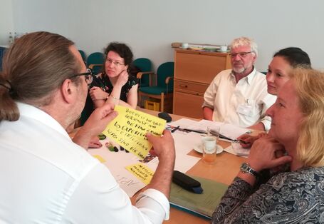 Teilnehmende sitzen am Gruppenarbeitstisch und hören einem Teilnehmenden zu. Er liest Arbeitsergebnisse von gelben Moderationskarten vor, die er in den Händen hält. Wir schauen ihm als Bildbetrachtende über die Schulter.