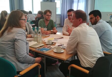 Teilnehmende sitzen im angeregten Gespräch miteinander am Gruppenarbeitstisch. Auf dem Tisch liegen Materialien wie Papier, Stifte, bunte Blätter. Es stehen Getränkeflaschen mit Wasser auf den Tischen sowie Tassen und Gläser.