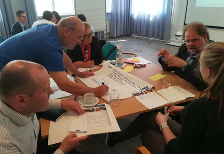 Teilnehmender beugt sich über den Gruppenarbeitstisch und schreibt auf Papier mit was ein Mitteilnehmender spricht.