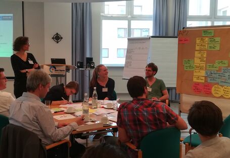 Teilnehmende sitzen in einer großen Runde am Gruppenarbeitstisch vor der Moderationswand mit den Arbeitsergebnisse auf farbigen Moderationskarten. Sie diskutieren angeregt und lachen miteinander.