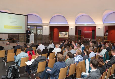 Viele Personen sitzen vor einer Bühne und hören einem Redner zu