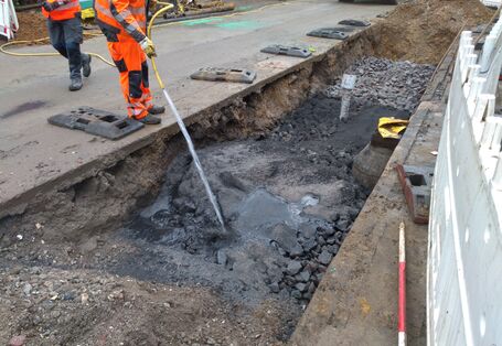 Fotografie einer Baustelle. In einem aufgerissenen Teilabschnitt der Straße wird ein dunkles Gemisch bewässert.