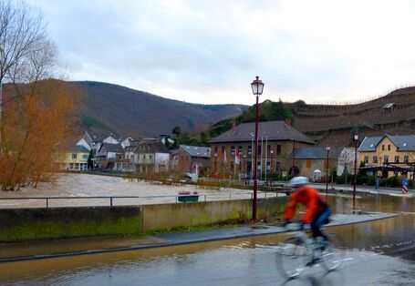 Person fährt mit dem Fahrrad durch Hochwasser in einer Ortschaft. Im Hintergrund sind Häuser und Berge zu sehen. Himmel ist bewölkt.