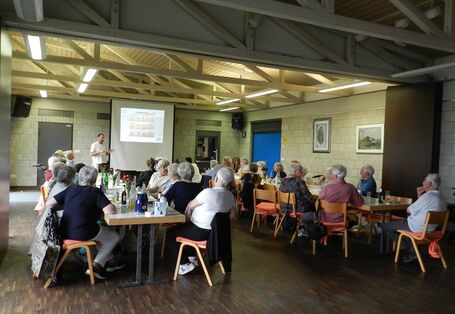 Bürger*innen sitzen im Vortragsraum an zwei langen Tischen parallel zu einander und schauen nach vorne auf die Präsentationsleinwand.