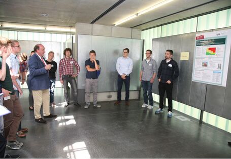 Studierende stehen kreisförmig um einen Dozenten. Im Hintergrund befinden sich silbernfarbene Stellwände.