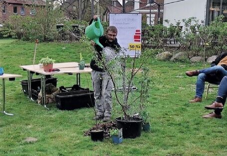 Der Veranstaltende gießt einen jungen Baum  mit einer grünen Gießkanne im Garten. Tische, Pflanzgefäße stehen im Hintergrund.