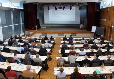 Von hinten oben wurde eine Halle fotografiert, in der an Tischen, die zu einer Bühne ausgerichtet ist, viele Menschen sitzen. Auf der Bühne hält ein Sprecher eine Präsentation.