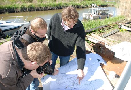 3 Männer stehen am Wasserlauf in der Sonne und schauen über eine auf dem Tisch ausgebreitete Karte. Ein Mann zeigt auf die Karte, wobei die anderen beiden Personen fotografieren und zusehen.